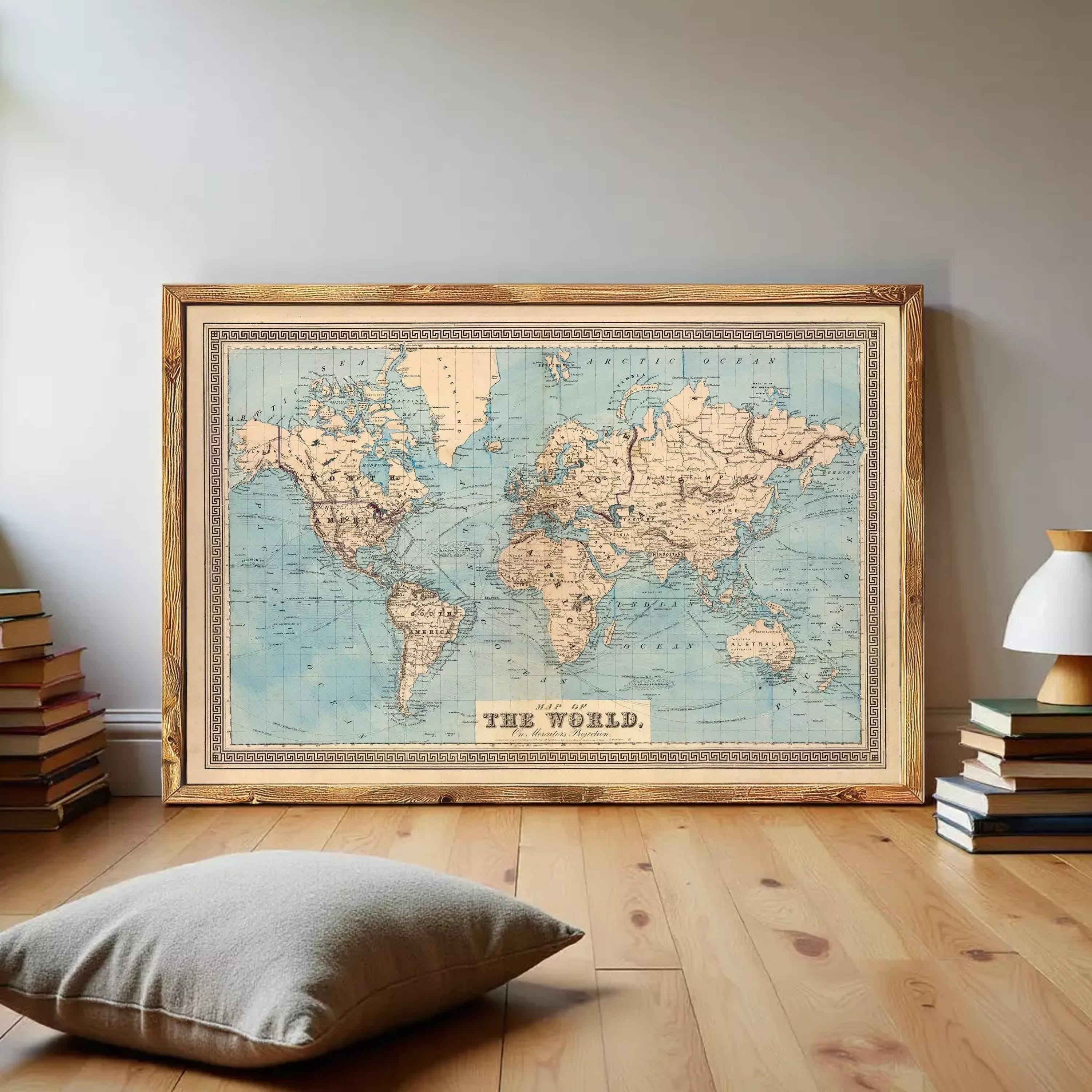 a large world map in a wooden frame, displayed on a wooden floor in a room with a stack of books and a pillow.